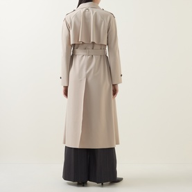 Model wearing HIGH Smuggler Trench Coat in beige, showing rear view with storm flap, matching belt, and longline silhouette over wide-leg trousers.