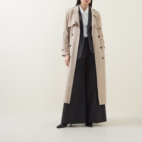 Model wearing HIGH Smuggler Trench Coat in beige, double-breasted longline style with belt, paired with black wide-leg trousers and heels.
