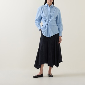 Model wearing HIGH Playful Tie Front Oversized Shirt in light blue with black asymmetric midi skirt and black flat shoes.