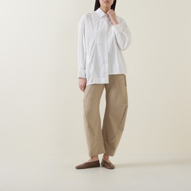 Model wearing HIGH Tumble Down Oversized Shirt in white with dropped shoulders and asymmetric hem, styled with beige tapered trousers and brown
