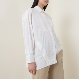 Model wearing HIGH Tumble Down Oversized Shirt in white cotton, with dropped shoulders, patch pocket, and asymmetric hem, styled with beige trousers.