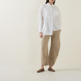 Model wearing HIGH Tumble Down Oversized Shirt in white with dropped shoulders and asymmetric hem, styled with beige wide-leg trousers and mules.