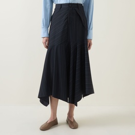 Model wearing HIGH Helix Asymmetric Pinstripe Skirt in navy, midi length with asymmetric hem, styled with a light blue shirt and taupe flats.