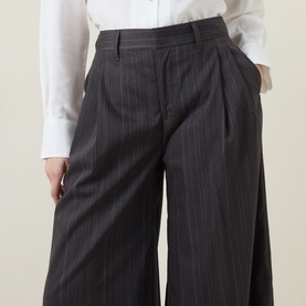 Model wearing HIGH Doff Pleated Pinstripe Trousers in dark grey; wide-leg, high-rise with front pleats, styled with a white shirt.