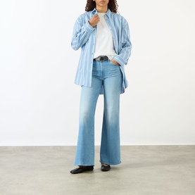 Model wearing Mother The Hustler High-Rise Wide Leg Jeans in Room Service light blue denim, styled with a blue striped oversized shirt and black belt.