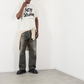 Model wearing Acne Gothic Logo T-Shirt in dusty white with dark wash wide-leg jeans, striped shirt tied at waist, and black loafers.