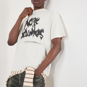 Model wearing Acne Gothic Logo T-Shirt in dusty white with black spray-painted gothic logo, styled with striped shirt and jeans.