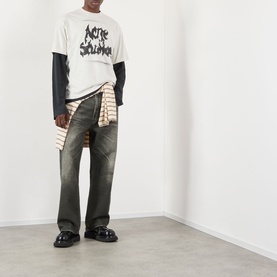 Model wearing Acne Gothic Logo T-Shirt in dusty white with dark layered long sleeves, styled with dark jeans and striped shirt tied at waist.
