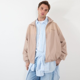 Model wearing Acne Washed Logo Jacket in mushroom beige with high collar, styled over a light blue striped shirt with tied sleeves.