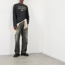 Model wearing Acne Sprayed 1996 Logo T-Shirt in faded black, long sleeve, styled with khaki wide-leg jeans and a striped shirt tied at waist.