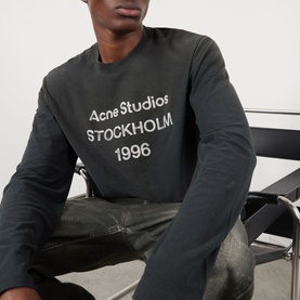Model wearing Acne Sprayed 1996 Logo T-Shirt in faded black, long sleeve crew neck with distressed stamp logo print, seated pose