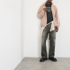 Model wearing Acne Sprayed 1996 Logo T-Shirt in faded black, styled with a pink zip-up jacket, striped shirt and khaki wide-leg jeans.