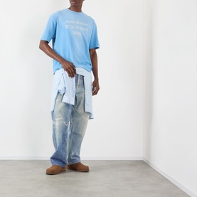 Model wearing Acne Sprayed Logo T-Shirt in cornflower blue with distressed wide-leg jeans and a shirt tied at the waist.