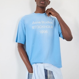 Model wearing Acne Sprayed Logo T-Shirt in cornflower blue with distressed stamp logo, styled with striped trousers tied at waist.