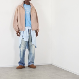 Model wearing Acne Sprayed Logo T-Shirt in cornflower blue, styled with a beige zip-up jacket, light blue shirt, distressed jeans, and tan suede