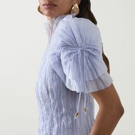 Model wearing Aje Clara Pintuck Dress in French Blue, showing pintuck detail, puff sleeve with bow and gold tassel, and high neckline