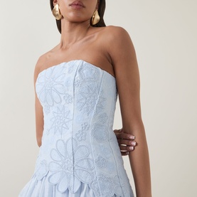 Model wearing Aje Gia Embroidered Mini Dress in Pastel Blue, strapless fitted bodice with mandala embroidery and peplum hem. Gold earrings visible.