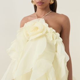 Model wearing Aje Pandorea Ruffle Dress in butter, halter neckline, layered floral ruffles, styled with gold hoop earrings