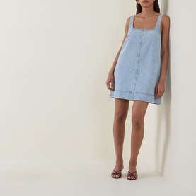 Model wearing Aje Lucina Studded Denim Mini Dress in light wash blue, square neck, sleeveless with stud detail, styled with burgundy strappy heels