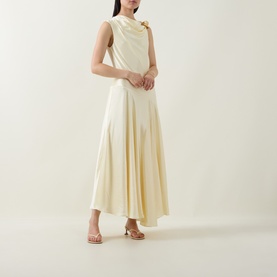 Model wearing Aje Siren Ruffled Dress in butter, sleeveless midi with asymmetric hem and gold-toned neckline hardware, styled with tan heeled sandals.
