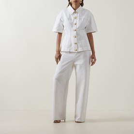 Model wearing Aje Liliana Denim Shirt in ivory with pointed collar, gold buttons, fitted waist and flared hem, styled with wide-leg white trousers.