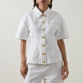 Model wearing Aje Liliana Denim Shirt in ivory with gold buttons, pointed collar, short sleeves, and fitted waist, styled with matching ivory