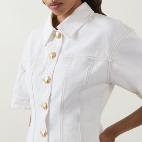 Model wearing Aje Liliana Denim Shirt in ivory with gold buttons, pointed collar, short sleeves, and fitted bodice with shaping seams.