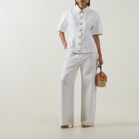 Model wearing Aje Liliana Denim Shirt in ivory with pointed collar, short sleeves and flared hem, styled with wide-leg white trousers and tan bag.