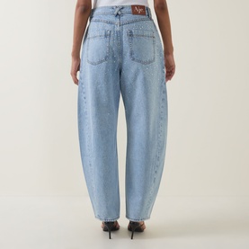 Model wearing Aje Lucina High-Rise Stud Barrel Jeans in light wash blue, rear view showing logo badge, tobacco stitching and barrel-leg silhouette.