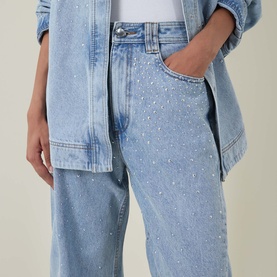 Model wearing Aje Lucina High-Rise Stud Barrel Jeans in light wash blue with crystal stud embellishment, styled with a denim shirt.