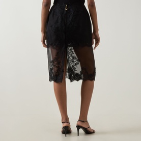 Model wearing Aje Ophelia Lace-Trim Skirt in black, showing sheer lace overlay and scalloped hem detail, styled with black strappy heels