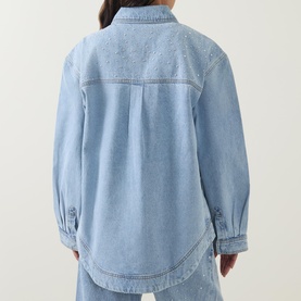 Model wearing Aje Lucina Denim Shirt in Light Wash Blue, rear view, showing dropped shoulders, long sleeves, and curved hem.