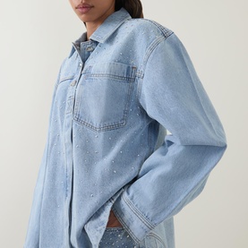 Model wearing Aje Lucina Denim Shirt in Light Wash Blue with raised stud detail, long sleeves, and snap-fastening cuffs.