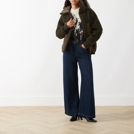 Model wearing HIGH olive faux shearling zip-up coat, styled with a printed top, wide-leg dark blue jeans and black heels.