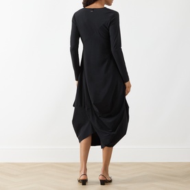 Model wearing HIGH black long-sleeve midi dress with draped asymmetric skirt, shown from behind, styled with black flat sandals.