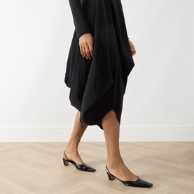 Model wearing HIGH black asymmetric draped dress with pointed-toe black slingback heels, shown from waist down