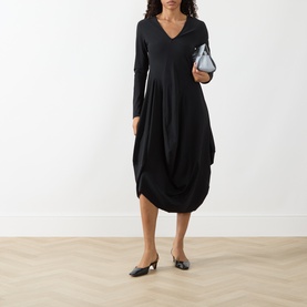 Model wearing HIGH black long-sleeve V-neck midi dress with draped hem, styled with black mules and a silver clutch bag.