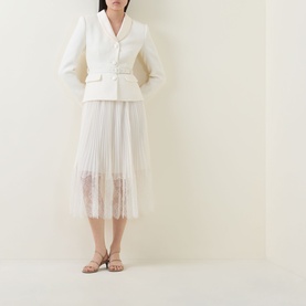Model wearing Self-Portrait cream dress with structured blazer-style bodice, pleated skirt with lace hem trim, styled with gold strappy heels