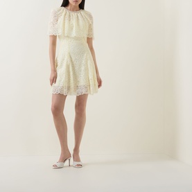 Model wearing Self-Portrait yellow lace mini dress with cape overlay and scalloped hem trim, styled with white heeled mules