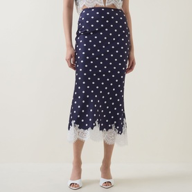 Model wearing Self-Portrait navy polka dot midi skirt with white lace hem trim, styled with white heeled mules and a white top.