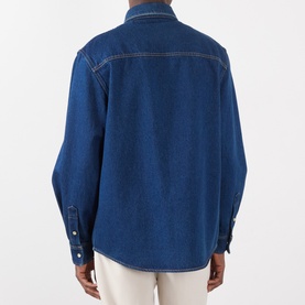 Model wearing Valentino medium blue denim cotton casual shirt, back view, with white contrast stitching and snap cuffs, styled with cream trousers.