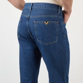 Model wearing Valentino medium blue denim jeans, rear view showing gold V logo hardware on back pocket, straight leg fit, 100% cotton