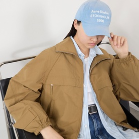 Model wearing Acne Logo Technical Bomber Jacket in dark khaki with blue logo cap, jeans and light blue shirt visible.