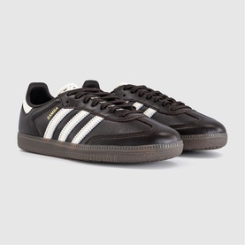 Adidas Samba trainers in black and off-white, featuring three white stripes, gold branding, black laces, and a dark gum sole.
