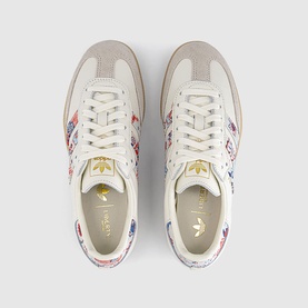 Adidas trainers in off-white with Liberty floral print detailing, gold bee emblem, cream suede panels, and gum sole, overhead view.