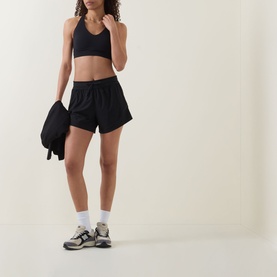 Model wearing black sports bra and black loose-fit shorts, styled with white socks, chunky trainers, and holding a black bag