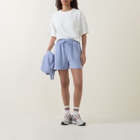 Model wearing light blue casual shorts and white tee, holding matching blue jacket, styled with white socks and chunky trainers