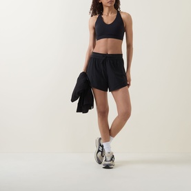 Model wearing black sports bra and black loose-fit shorts, carrying a black jacket, styled with white trainers and socks