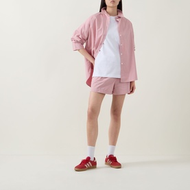 Model wearing Sweaty Betty Holiday Graphic T-Shirt in white, styled with pink oversized shirt, matching shorts and red Adidas Gazelle trainers.