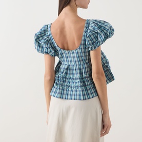 Model wearing Damson Madder blue check cotton blouse with puffed sleeves, low scoop back, and ruched waist detail, styled with cream wide-leg